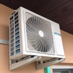 Biała jednostka zewnętrzna klimatyzacji marki HEIKO zamontowana na wspornikach na elewacji budynku z drewnianym okapem.