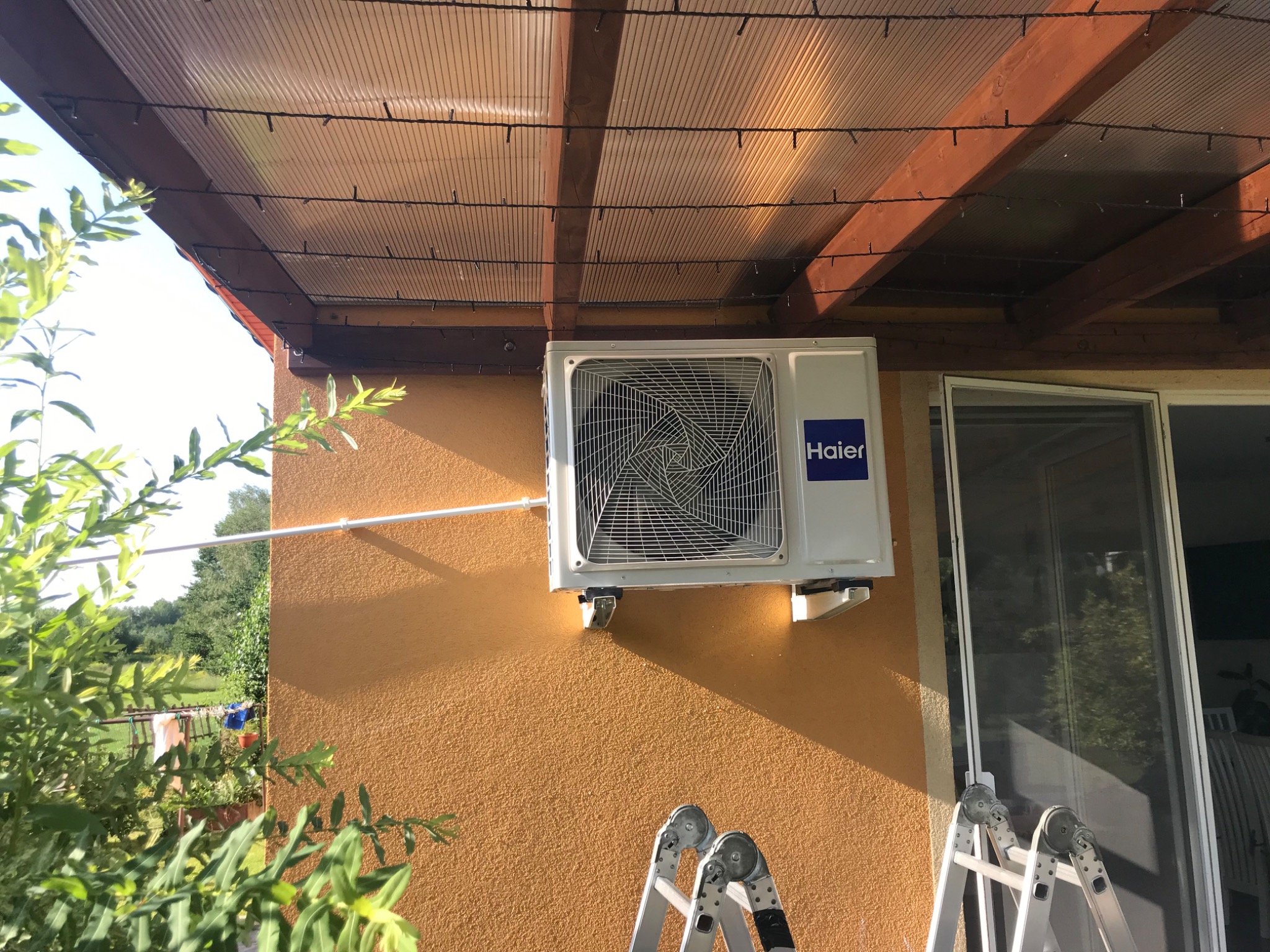Zewnętrzna jednostka klimatyzacji Haier zamontowana na pomarańczowej elewacji domu, widoczne drabiny aluminiowe, zadaszenie z poliwęglanu z lampkami oraz fragment ogrodu.