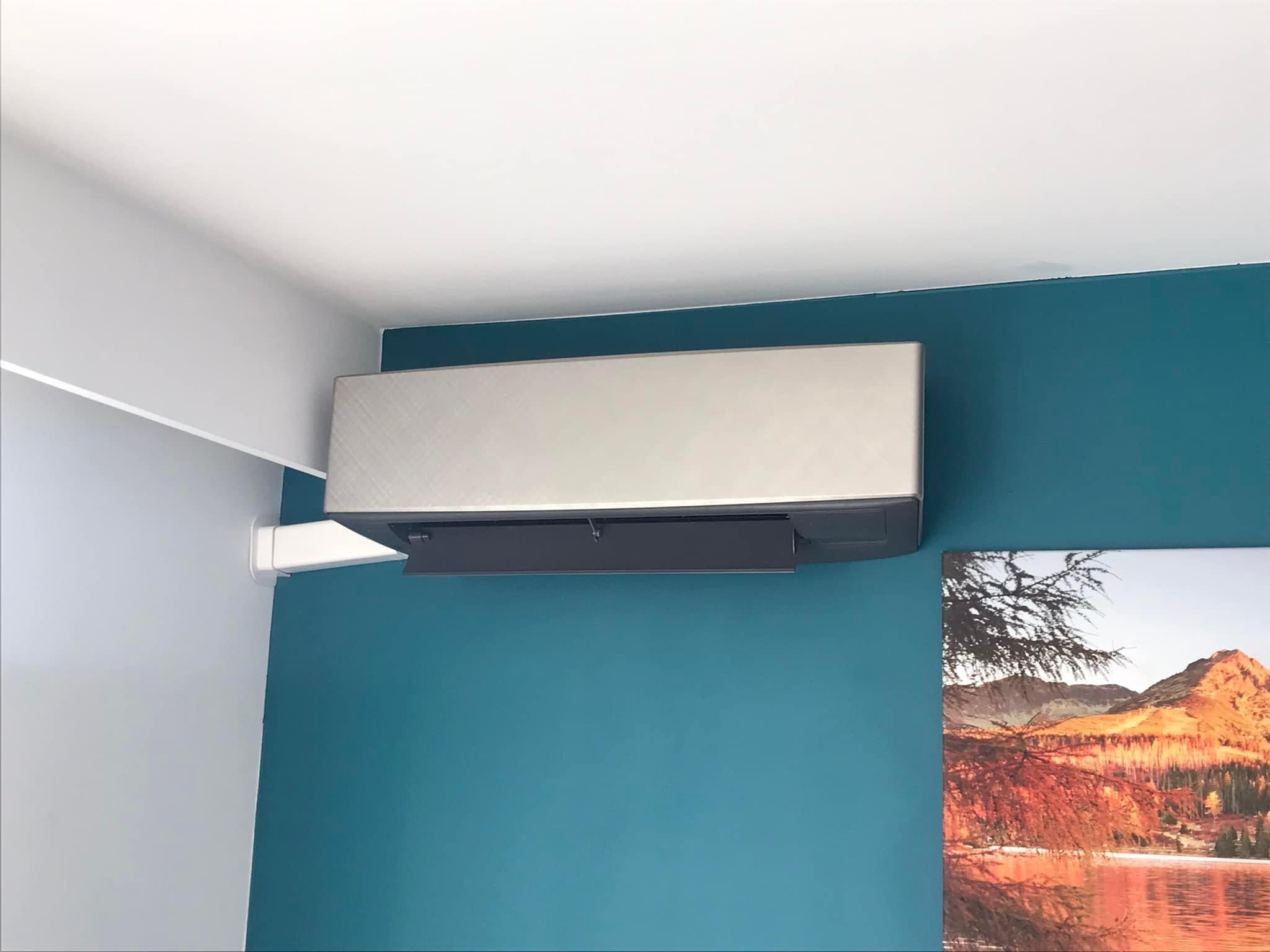 Klimatyzator ścienny z srebrnym panelem, zamontowany w rogu pokoju pomalowanego na turkusowo, obok wisi obraz z górskim krajobrazem.