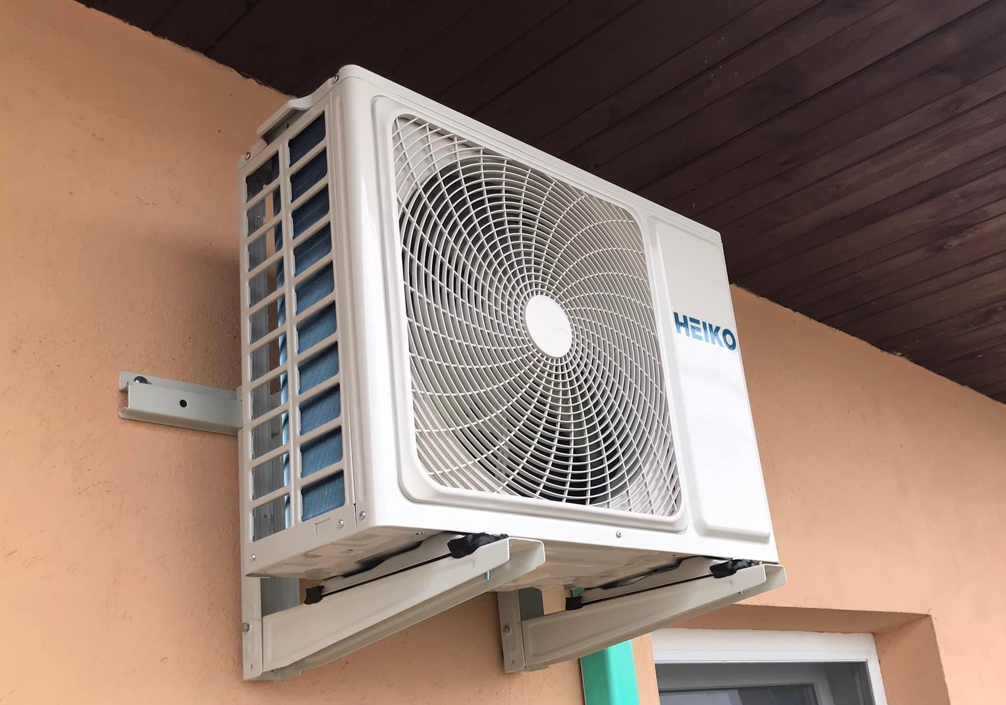 Biała jednostka zewnętrzna klimatyzacji marki HEIKO zamontowana na wspornikach na elewacji budynku z drewnianym okapem.