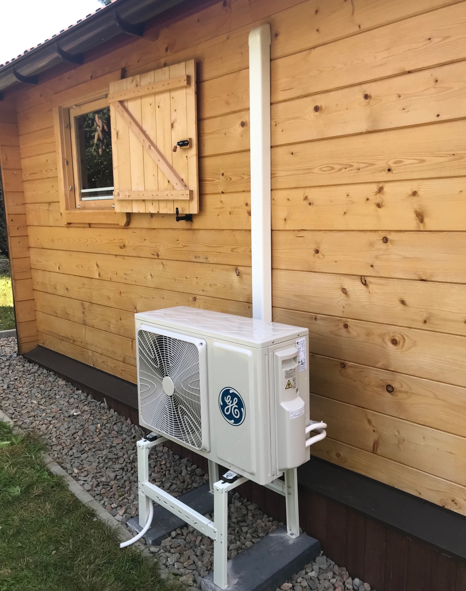 Zewnętrzna jednostka klimatyzacji marki GE zamontowana na białym stelażu przy drewnianej ścianie budynku z okiennicą.
