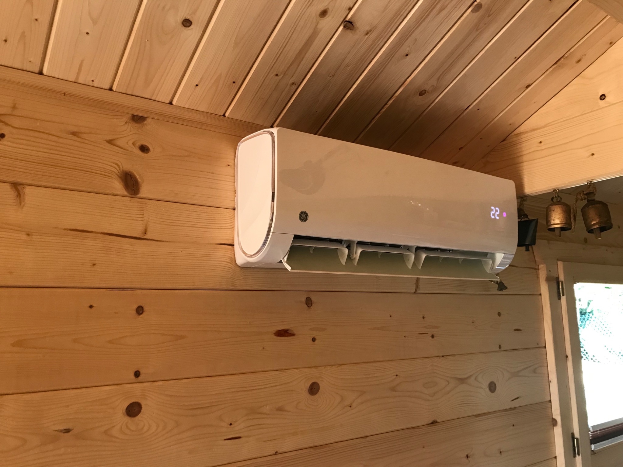 Biała jednostka klimatyzacji ściennej marki GE zamontowana w drewnianym domku, widoczny wyświetlacz wskazujący 22 stopnie, obok okno z brązowymi dzwoneczkami.