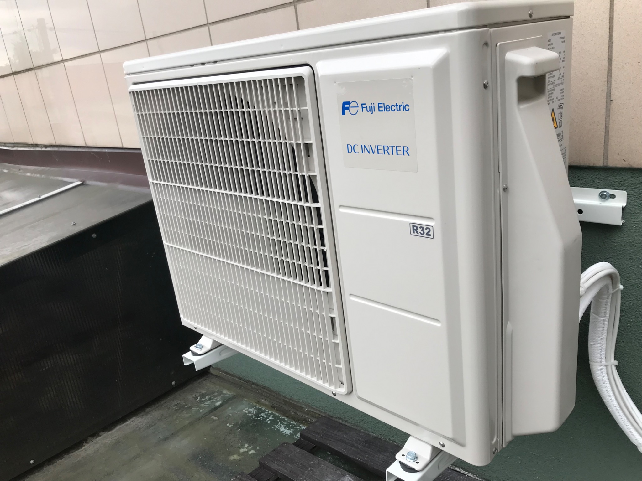 Biała zewnętrzna jednostka klimatyzacji Fuji Electric DC Inverter R32 zamontowana na zewnątrz budynku, widoczne przewody chłodnicze.