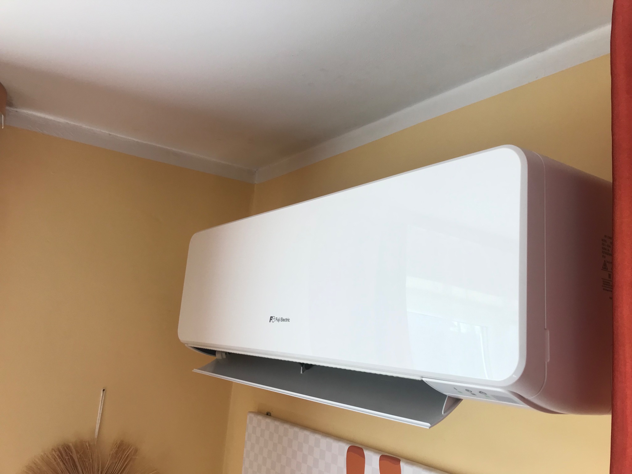 Biała klimatyzacja Fuji Electric zamontowana na żółtej ścianie w pomieszczeniu z widocznym fragmentem zasłony i dekoracyjnym wachlarzem.
