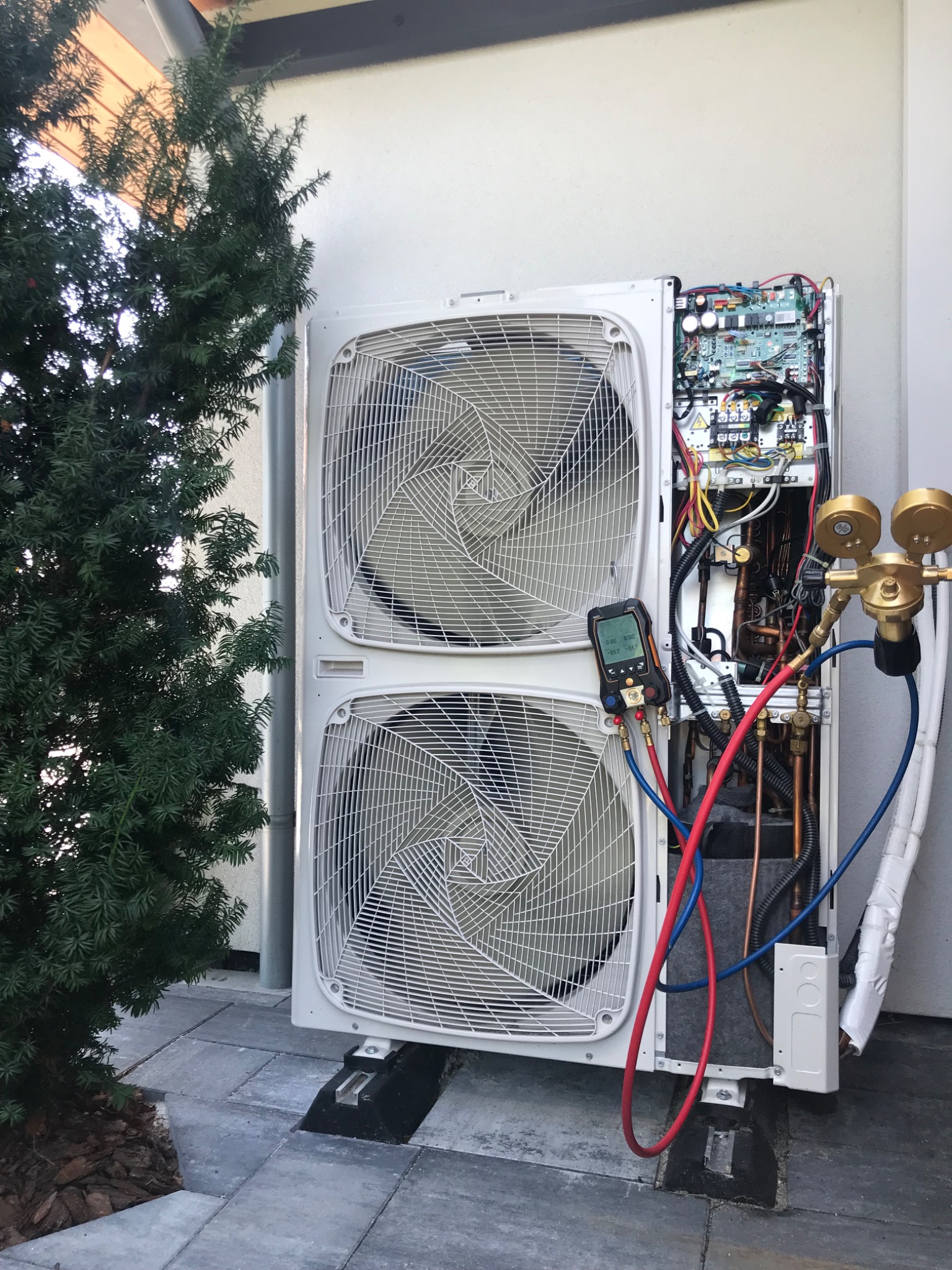 Zewnętrzna jednostka klimatyzacji z otwartym panelem bocznym, podłączonymi przewodami pomiarowymi i manometrami, stojąca na betonowej kostce brukowej obok iglastego krzewu.