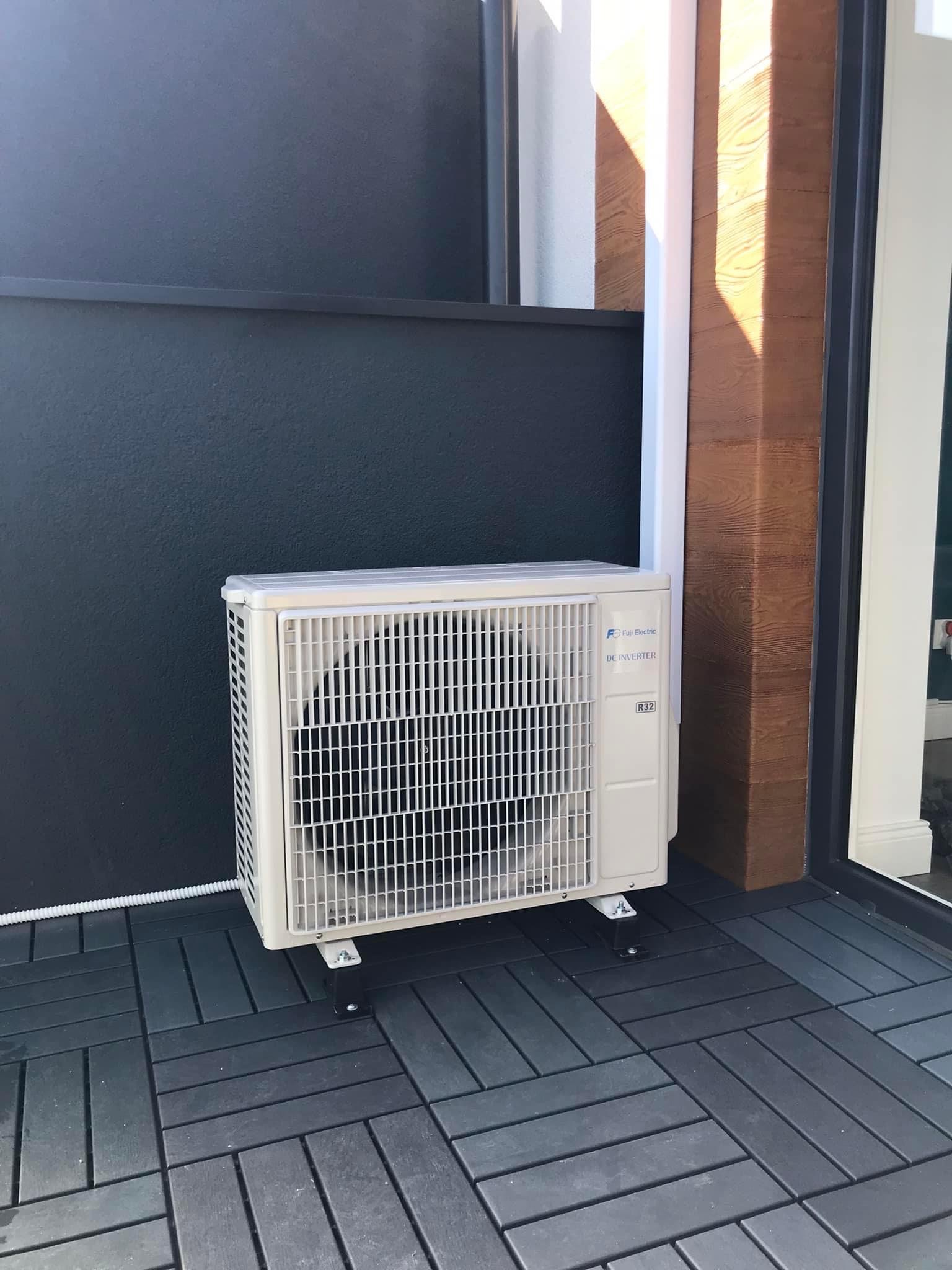 Zewnętrzna jednostka klimatyzacji Fuji Electric DC Inverter R32, zamontowana na czarnych wspornikach na szarej, kwadratowej podłodze, przy ciemnej ścianie z drewnianym wykończeniem.
