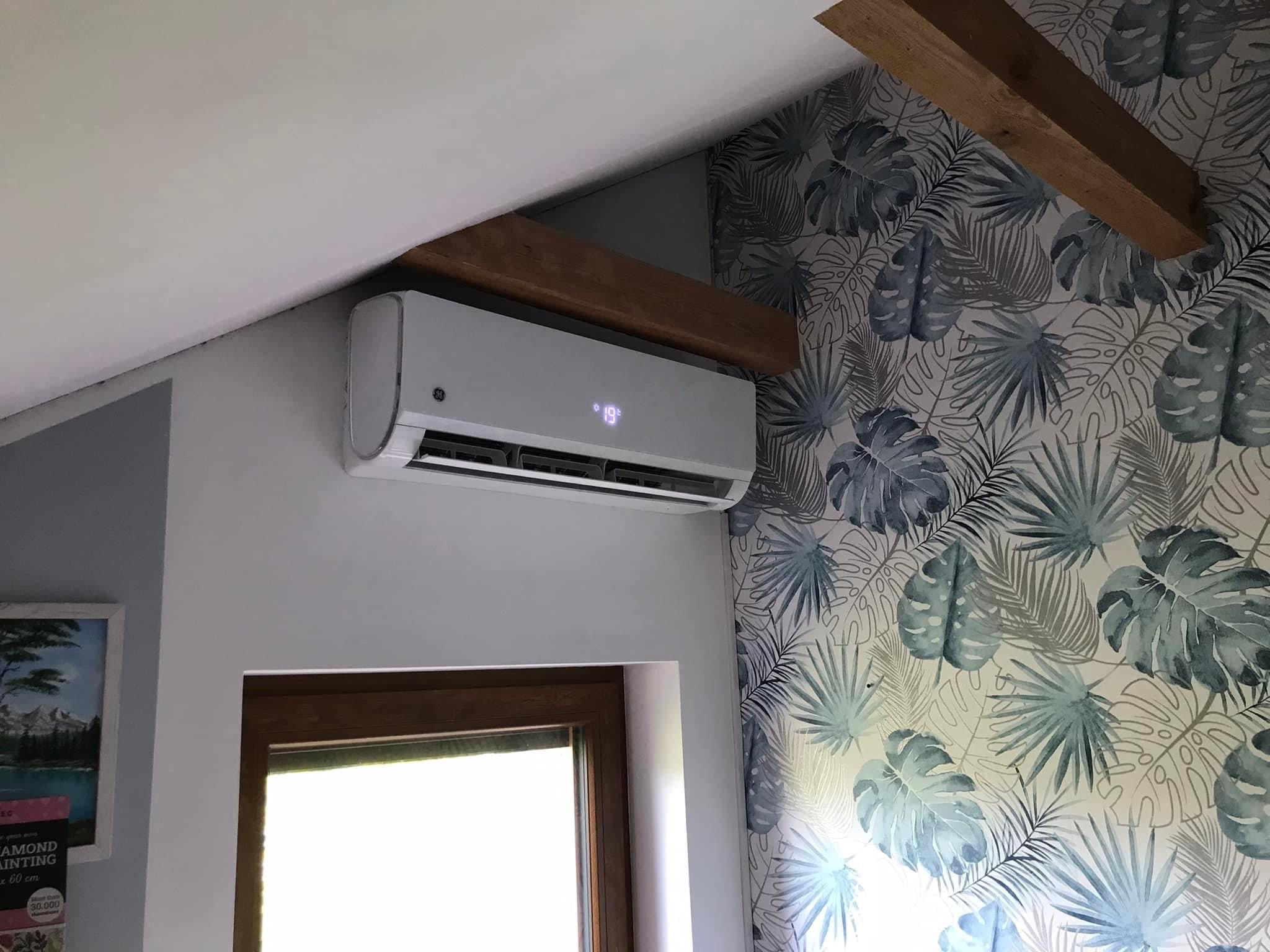 Biała jednostka klimatyzacji ściennej marki GE, zamontowana w pomieszczeniu z drewnianą belką stropową i tapetą w motywy roślinne, wyświetlająca temperaturę 19 stopni Celsjusza.