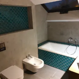 Nowoczesna łazienka z betonowymi ścianami, turkusową mozaiką w kształcie rybiej łuski na obudowie wanny i wnęce ściennej, białą armaturą (toaleta, bidet, wanna) oraz oknem dachowym.