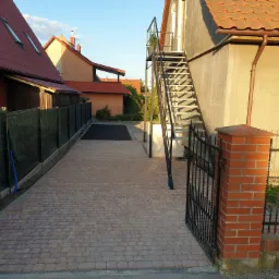 Ułożona kostka brukowa na podjeździe, czarne metalowe ogrodzenie z ceglanym słupkiem, metalowe schody zewnętrzne do budynku, domy z czerwonymi dachami w tle, błękitne niebo.