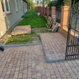 Ułożona kostka brukowa w odcieniach beżu i brązu prowadząca do furtki z metaloplastyki, widoczne palety i cegły przygotowane do dalszych prac.