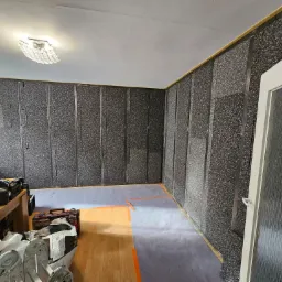 Pokój w trakcie prac wykończeniowych, ściany pokryte panelami izolacyjnymi o szorstkiej fakturze, widoczne metalowe profile montażowe, podłoga częściowo zabezpieczona folią, narzędzia i skrzynki...