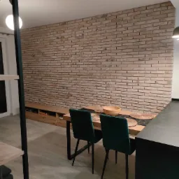 Jasna ceglana ściana w salonie z drewnianym stołem, zielonymi krzesłami i czarnym blatem kuchennym, oświetlona wiszącymi lampami