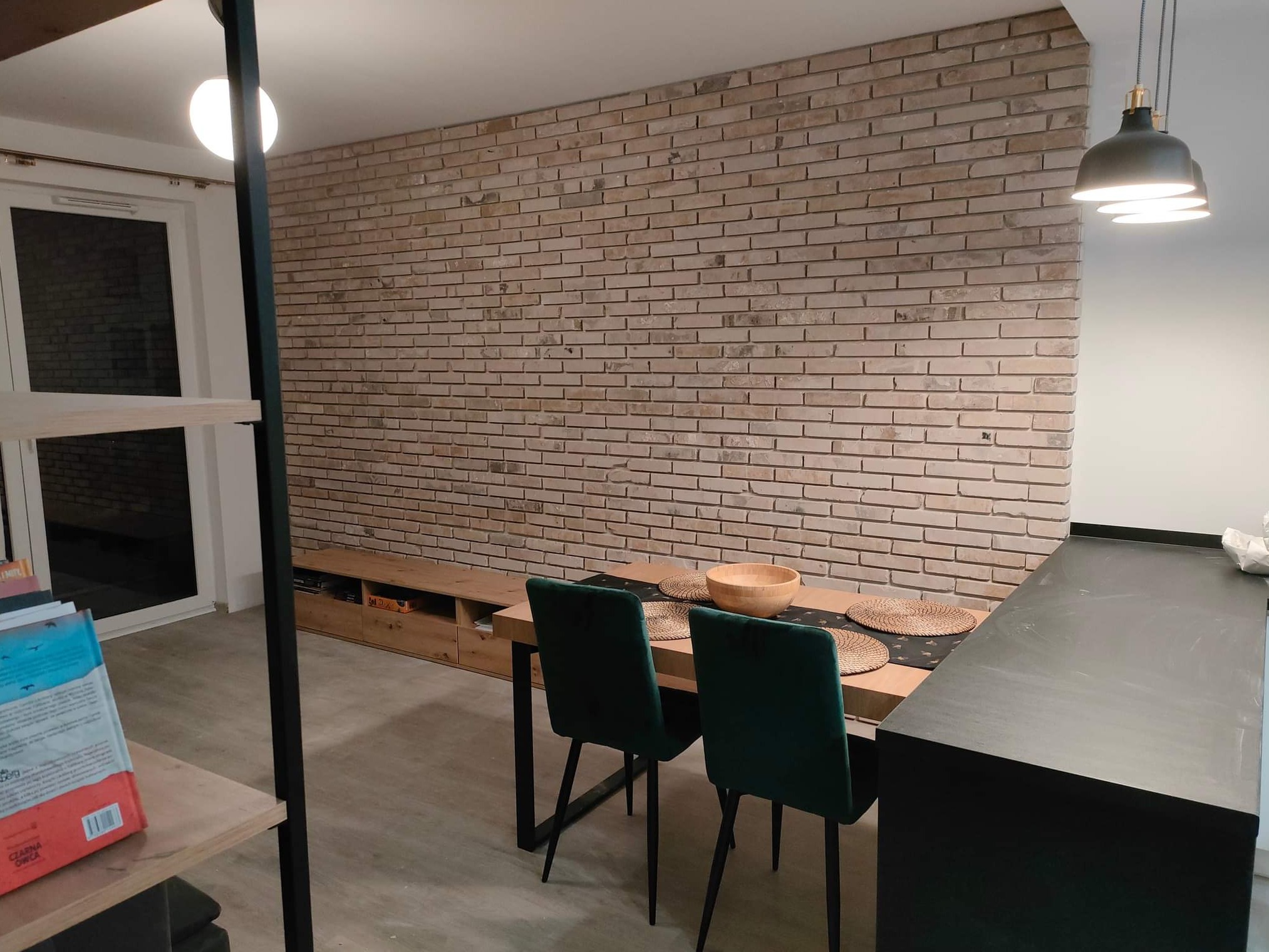 Jasna ceglana ściana w salonie z drewnianym stołem, zielonymi krzesłami i czarnym blatem kuchennym, oświetlona wiszącymi lampami