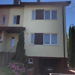 Malcobud - Dwukondygnacyjny dom z elewacją w odcieniach beżu, z brązowymi akcentami na oknach i dachu, garażem w przyziemiu i kwitnącymi hortensjami w ogrodzie.