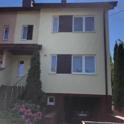 Dwukondygnacyjny dom z elewacją w odcieniach beżu, z brązowymi akcentami na oknach i dachu, garażem w przyziemiu i kwitnącymi hortensjami w ogrodzie.