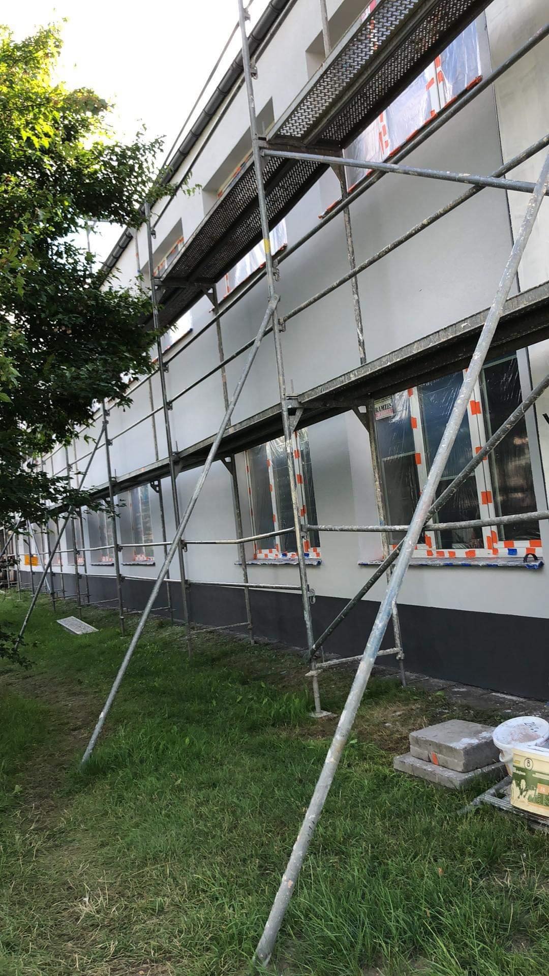 Budynek w trakcie prac elewacyjnych: Ściany otynkowane na biało, okna zabezpieczone folią, rusztowanie ustawione wzdłuż ściany, widoczne materiały budowlane na trawniku.