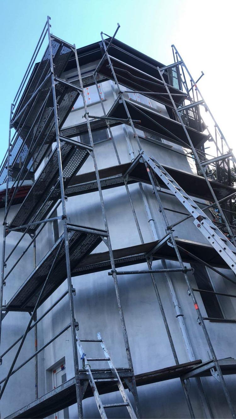 Budynek w trakcie prac elewacyjnych, widoczne rusztowania i ściana pokryta szarą warstwą wykończeniową, drabina oparta o rusztowanie, błękitne niebo w tle.