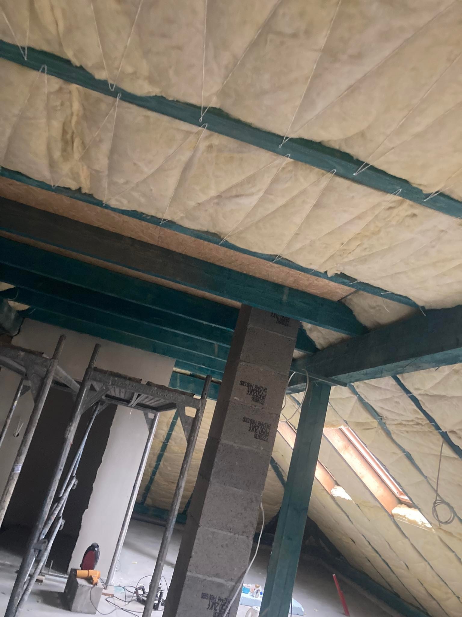 Izolacja poddasza wełną mineralną pomiędzy krokwiami, widoczne rusztowanie, okno dachowe i narzędzia.