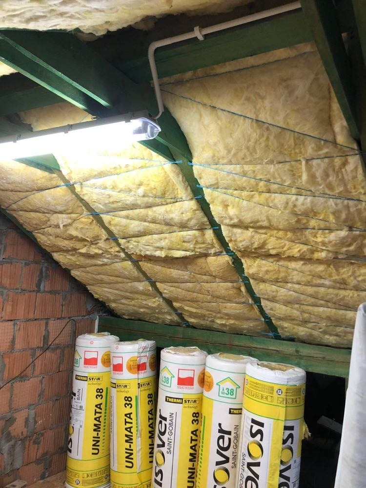 Izolacja poddasza wełną mineralną Isover UNI-MATA 38, widoczne rolki materiału i zamocowana izolacja między krokwiami, oświetlenie lampą jarzeniową.