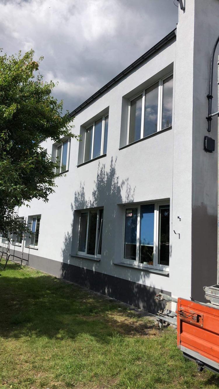 Dwu kondygnacyjny budynek po odnowieniu elewacji, z widocznymi oknami odbijającymi niebo, drzewem rzucającym cień na ścianę i przyczepką na trawniku.