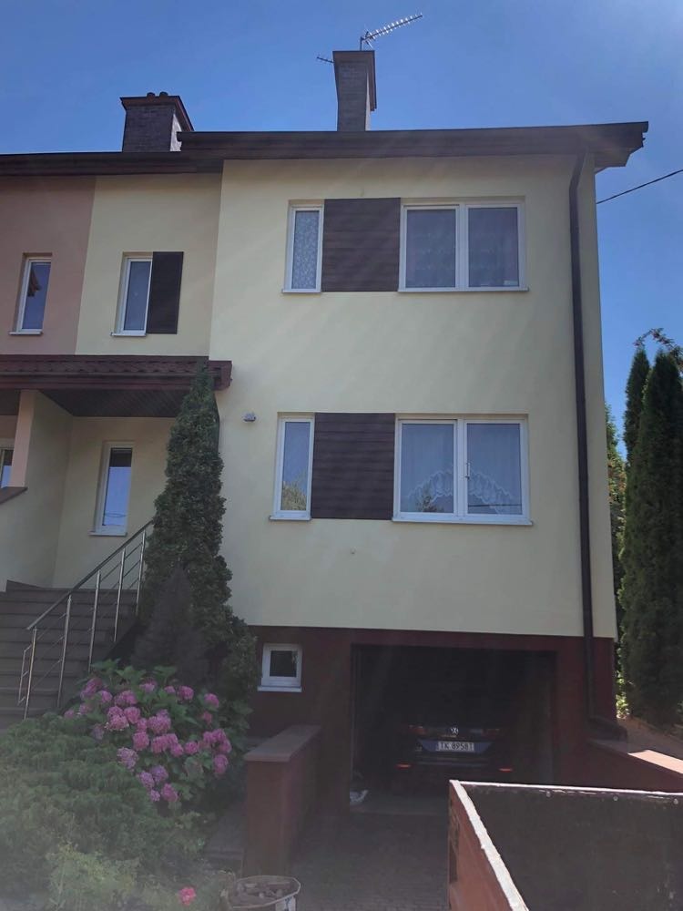 Dwukondygnacyjny dom z elewacją w odcieniach beżu, z brązowymi akcentami na oknach i dachu, garażem w przyziemiu i kwitnącymi hortensjami w ogrodzie.