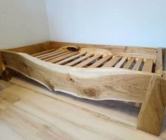 Łóżko z litego drewna o nieregularnym kształcie krawędzi, z widoczną naturalną strukturą słojów i minimalistycznym zagłówkiem z otworem. Stelaż listwowy. Podłoga drewniana w tle.