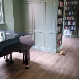 Tomasz Gorski - Wnętrze z zabudową w kolorze szałwii: szafa wnękowa połączona z regałem na książki, fortepian z czarnym połyskiem, panele podłogowe imitujące drewno dębowe.