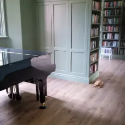 Wnętrze z zabudową w kolorze szałwii: szafa wnękowa połączona z regałem na książki, fortepian z czarnym połyskiem, panele podłogowe imitujące drewno dębowe.