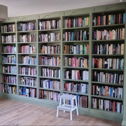 Tomasz Gorski - Wbudowany regał na książki w kolorze pistacjowym, wypełniony książkami, z małym białym stołkiem drabinkowym na drewnianej podłodze, widoczne oświetlenie punktowe nad półkami.