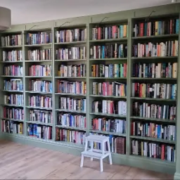 Wbudowany regał na książki w kolorze pistacjowym, wypełniony książkami, z małym białym stołkiem drabinkowym na drewnianej podłodze, widoczne oświetlenie punktowe nad półkami.