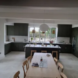 Przestronna kuchnia z ciemnozielonymi szafkami, białym blatem i wyspą, oświetlona wiszącymi lampami, z drewnianym stołem i krzesłami na pierwszym planie.