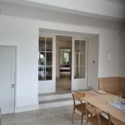 Jasne wnętrze z drewnianym stołem i krzesłami, widok na salon przez dwuskrzydłowe, przeszklone drzwi, kamienna podłoga i białe ściany.