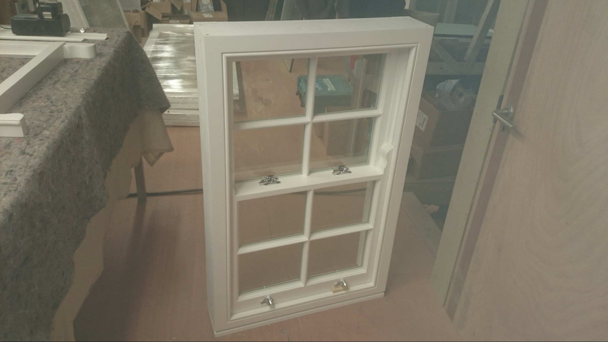 Białe, drewniane okno skrzynkowe z sześcioma szybami, widoczne okucia, w tle elementy warsztatu.