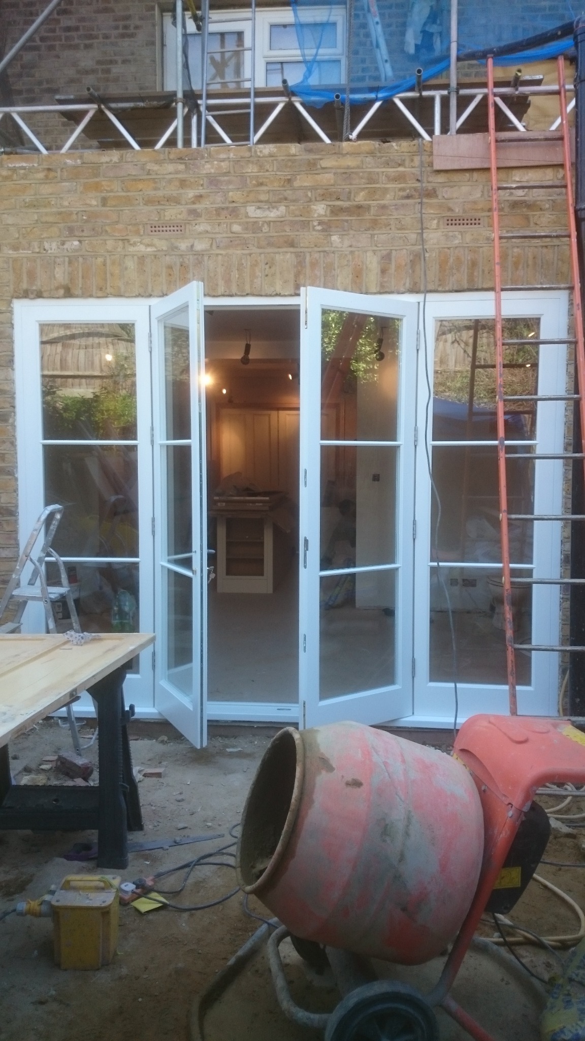 Montaż białych, przeszklonych drzwi balkonowych w trakcie prac wykończeniowych; widoczna ceglana ściana, rusztowanie, drabina oraz betoniarka.