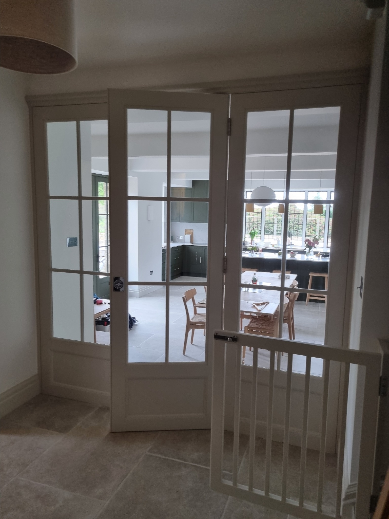 Białe, przeszklone drzwi wewnętrzne z widokiem na kuchnię i jadalnię; w tle widoczna wyspa kuchenna i stół z krzesłami.