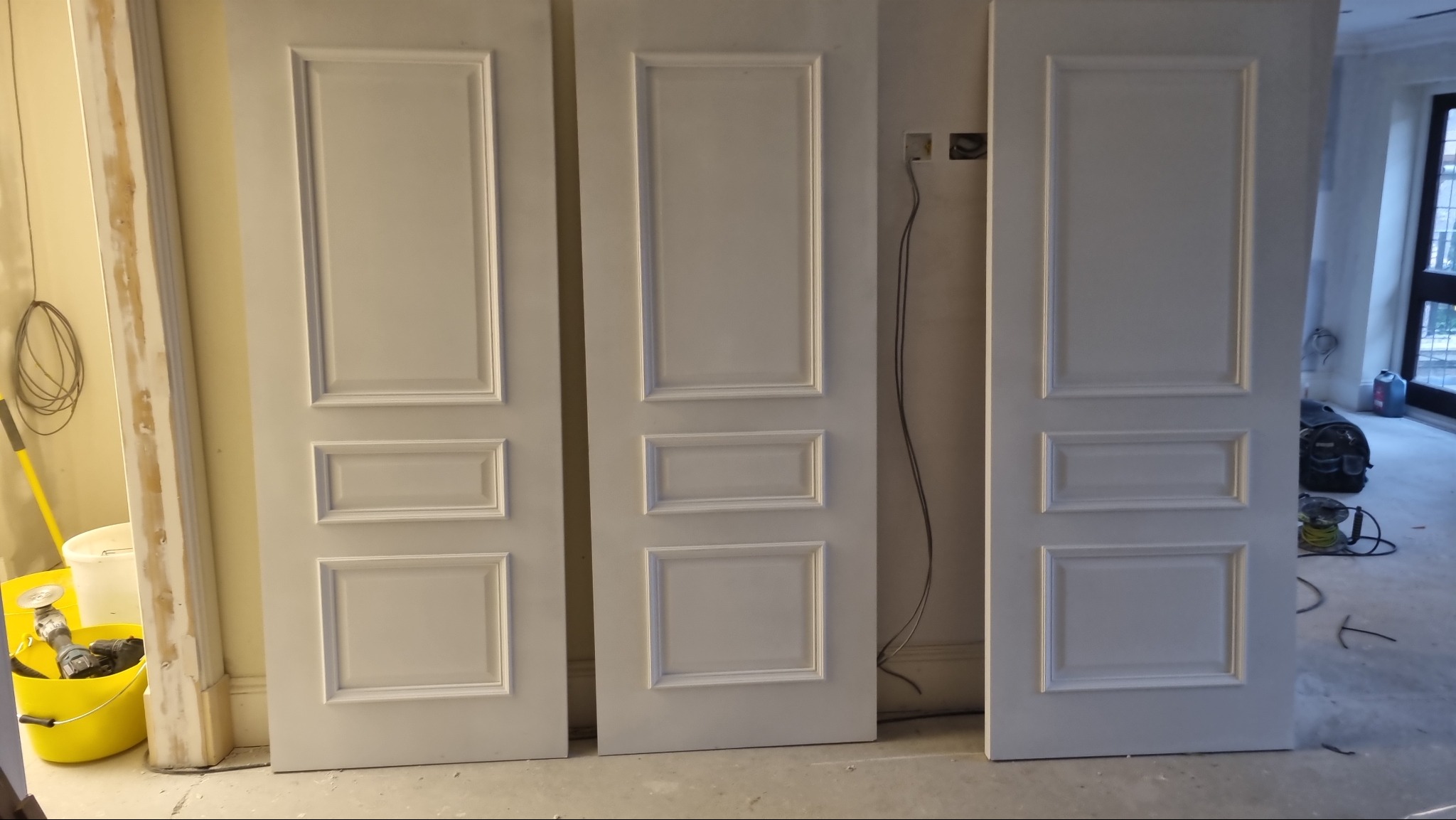 Trzy białe, panelowe drzwi wewnętrzne ustawione obok siebie w trakcie montażu, widoczne narzędzia i wiadro z akcesoriami, surowy stan wykończenia wnętrza.
