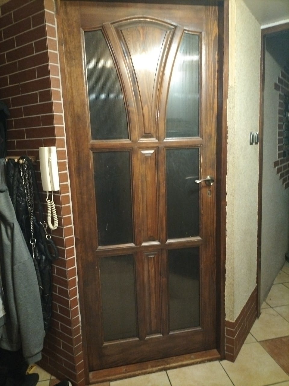 Drewniane drzwi wewnętrzne z szybami, widoczne z bliska, osadzone w ścianie wyłożonej cegłą, z fragmentem wieszaka na ubrania po lewej stronie.