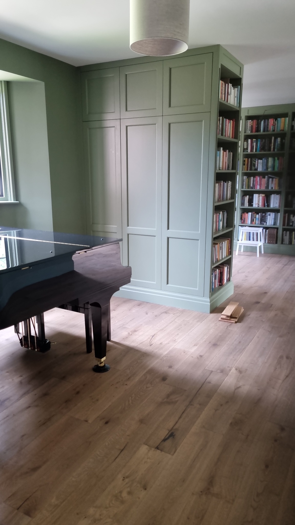 Wnętrze z zabudową w kolorze szałwii: szafa wnękowa połączona z regałem na książki, fortepian z czarnym połyskiem, panele podłogowe imitujące drewno dębowe.