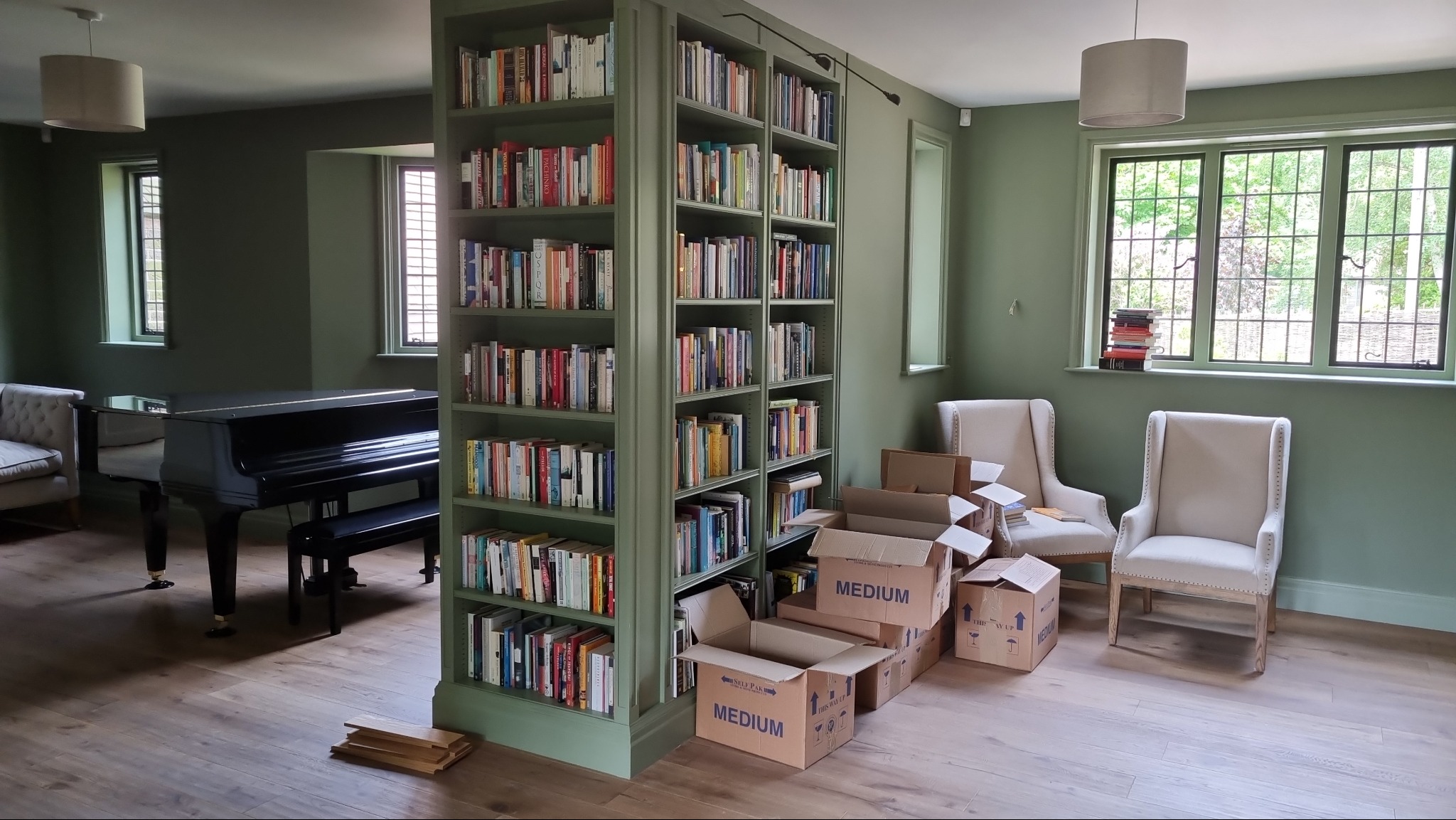 Wnętrze pokoju z zielonymi ścianami, zabudowaną biblioteczką z książkami, czarnym fortepianem i krzesłami, kartony do pakowania sugerują przeprowadzkę lub remont.