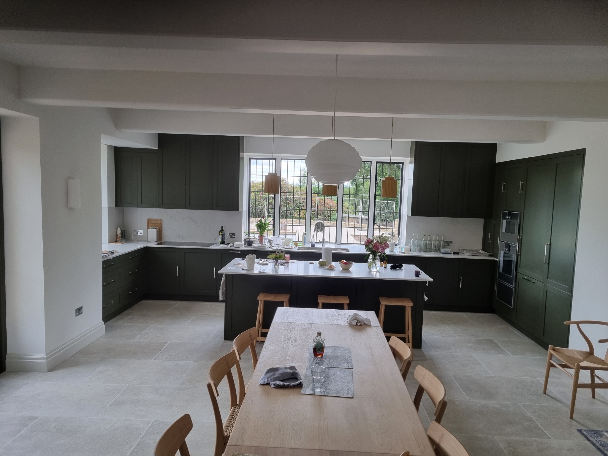 Przestronna kuchnia z ciemnozielonymi szafkami, białym blatem i wyspą, oświetlona wiszącymi lampami, z drewnianym stołem i krzesłami na pierwszym planie.