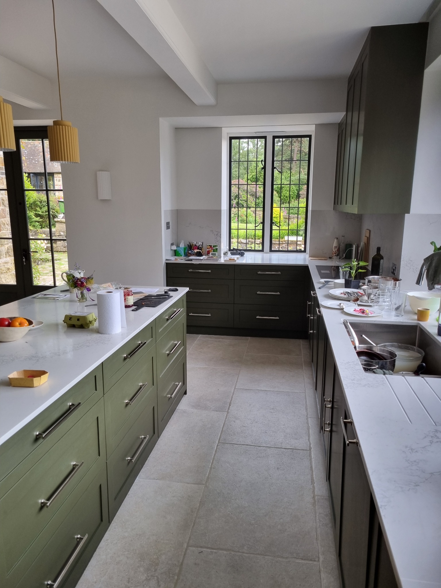 Wnętrze kuchni w odcieniach zieleni z kamienną podłogą, wyspą kuchenną z blatem z jasnego marmuru, zlewozmywakiem pełnym naczyń i widokiem na ogród przez okno.