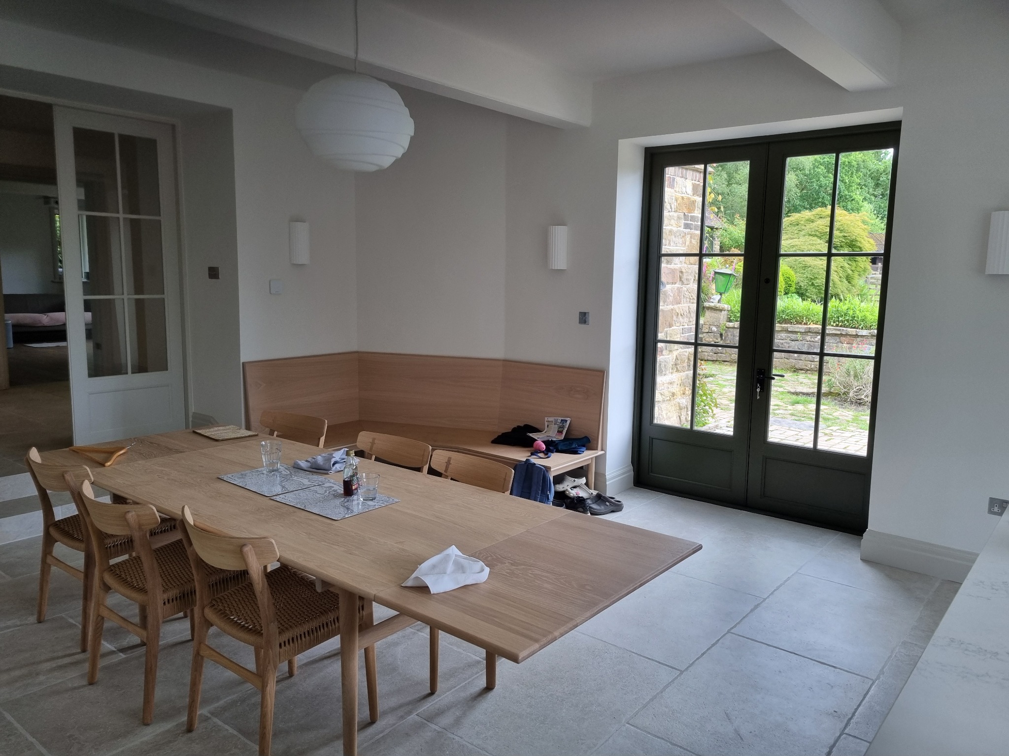 Jasna jadalnia z drewnianym stołem, krzesłami z plecionki, wbudowaną ławką i zielonymi, przeszklonymi drzwiami prowadzącymi do ogrodu