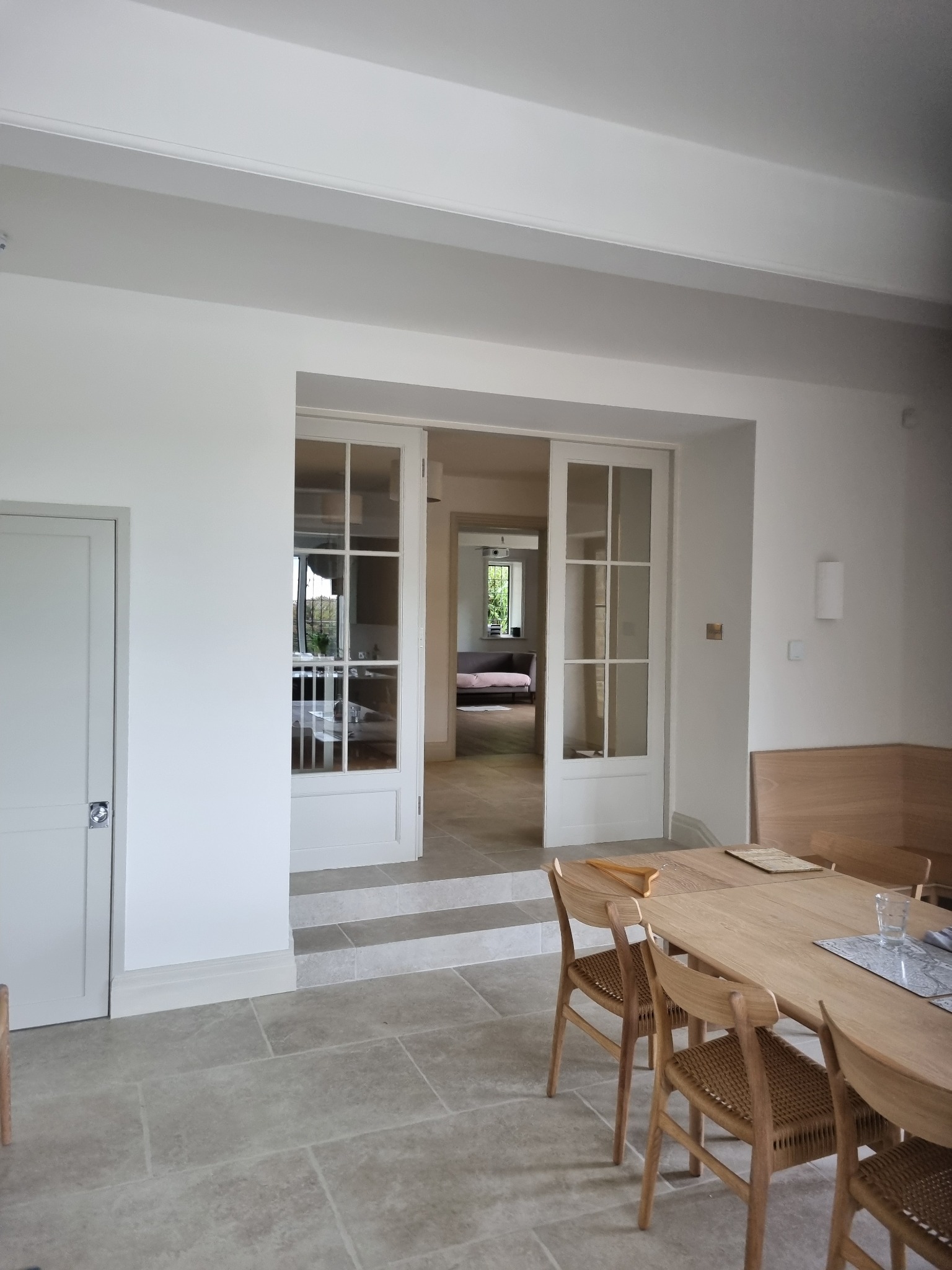 Jasne wnętrze z drewnianym stołem i krzesłami, widok na salon przez dwuskrzydłowe, przeszklone drzwi, kamienna podłoga i białe ściany.