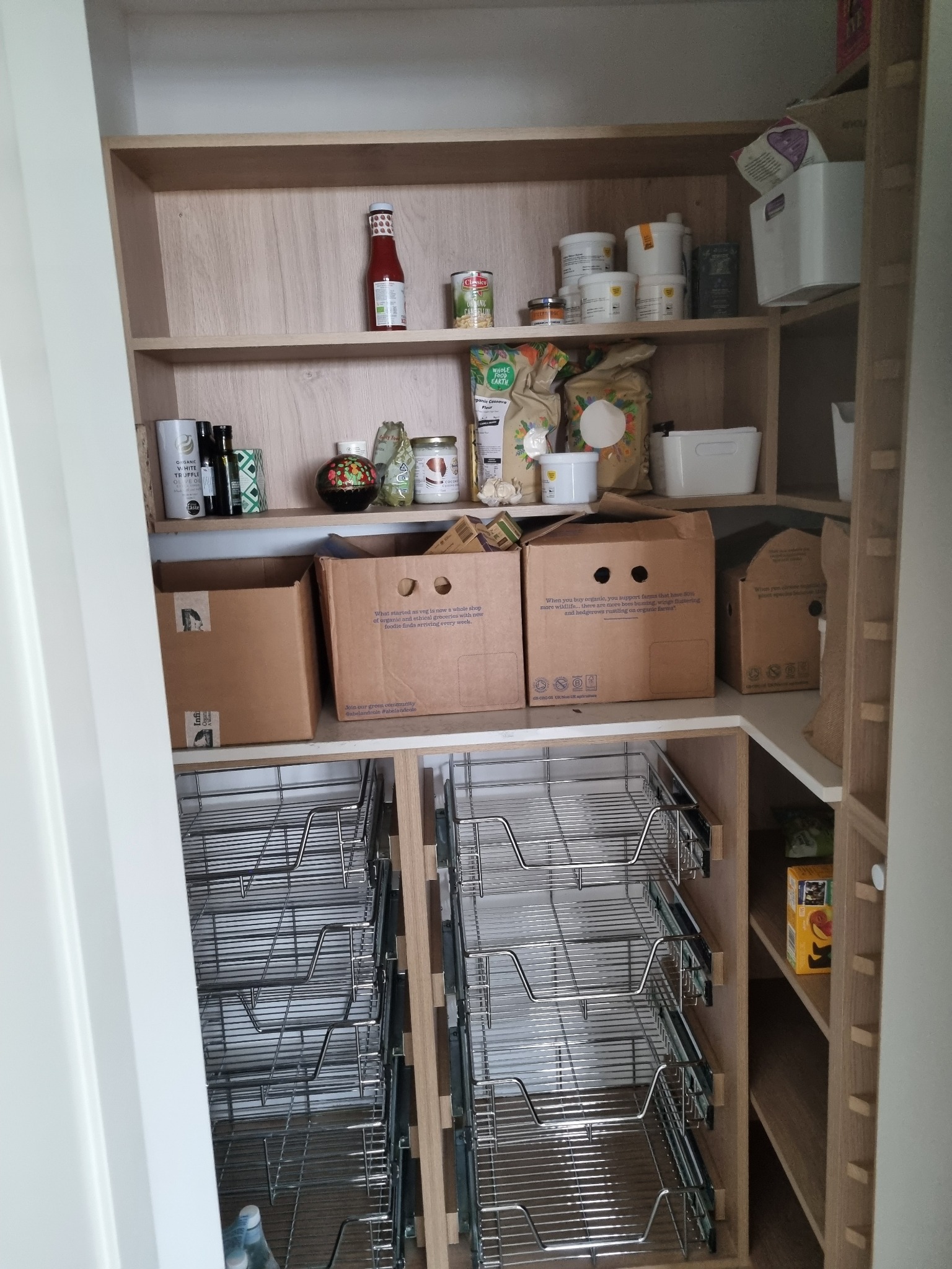 Wnętrze spiżarni z drewnianymi półkami i wysuwanymi metalowymi koszami. Na półkach widoczne zapasy żywności w słoikach, puszkach i kartonach. Dostęp do spiżarni przez białe drzwi po lewej stronie.