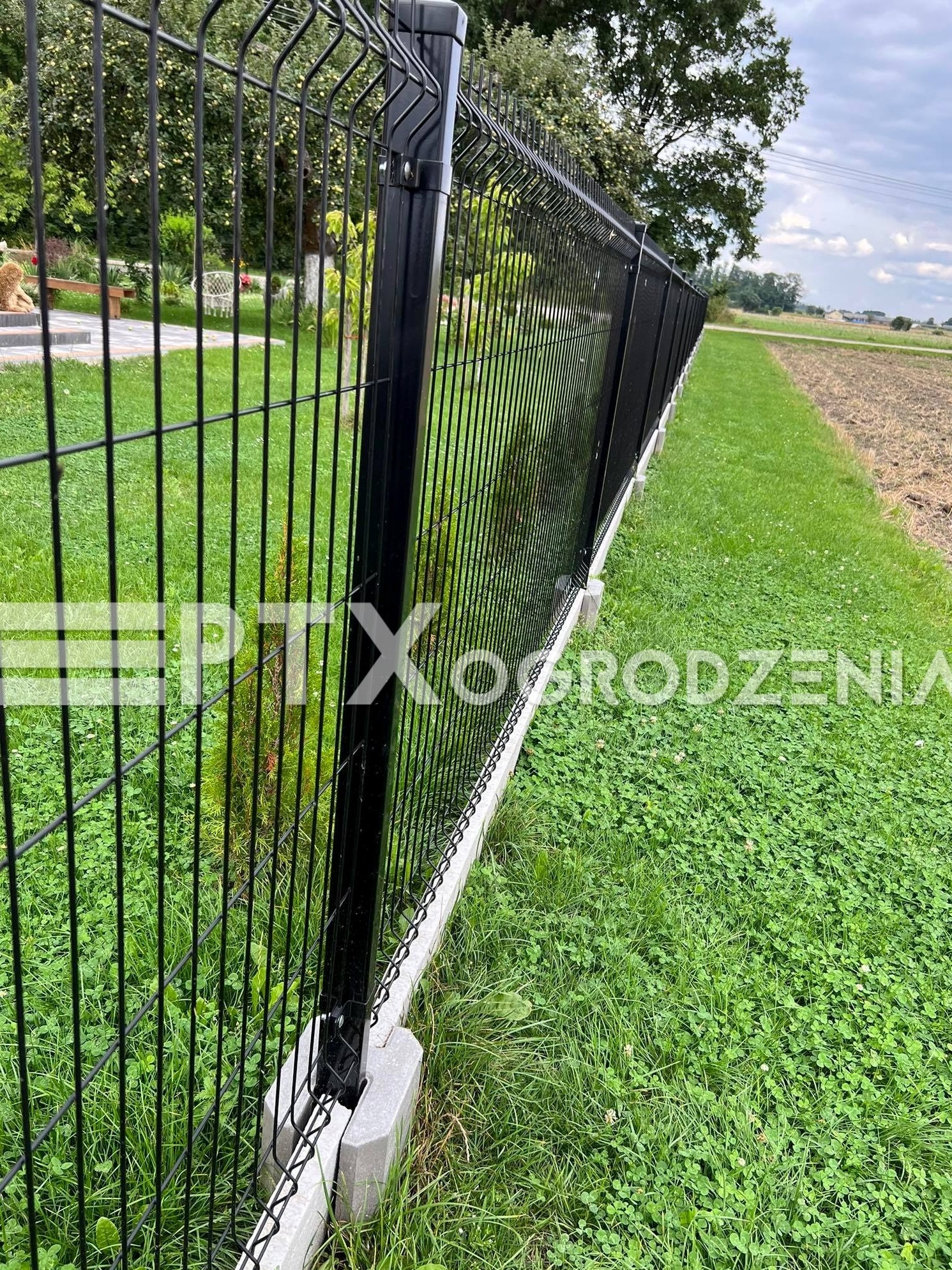 Czarny panelowy system ogrodzeniowy z podmurówką betonową, widok z perspektywy trawnika porośniętego koniczyną, w tle zieleń i pole uprawne.