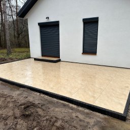 M.K.Bruk - Jasna, kwadratowa kostka na tarasie przy białym domu z szarymi roletami. Czarny obrzeże kostki. Widoczne drzewa w tle. Ukończony projekt.