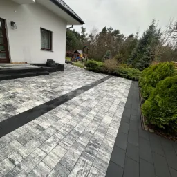 Nowoczesny podjazd z kostki brukowej w odcieniach szarości, z czarnymi pasami i obrzeżami, prowadzący do domu z widocznym placem zabaw w tle.