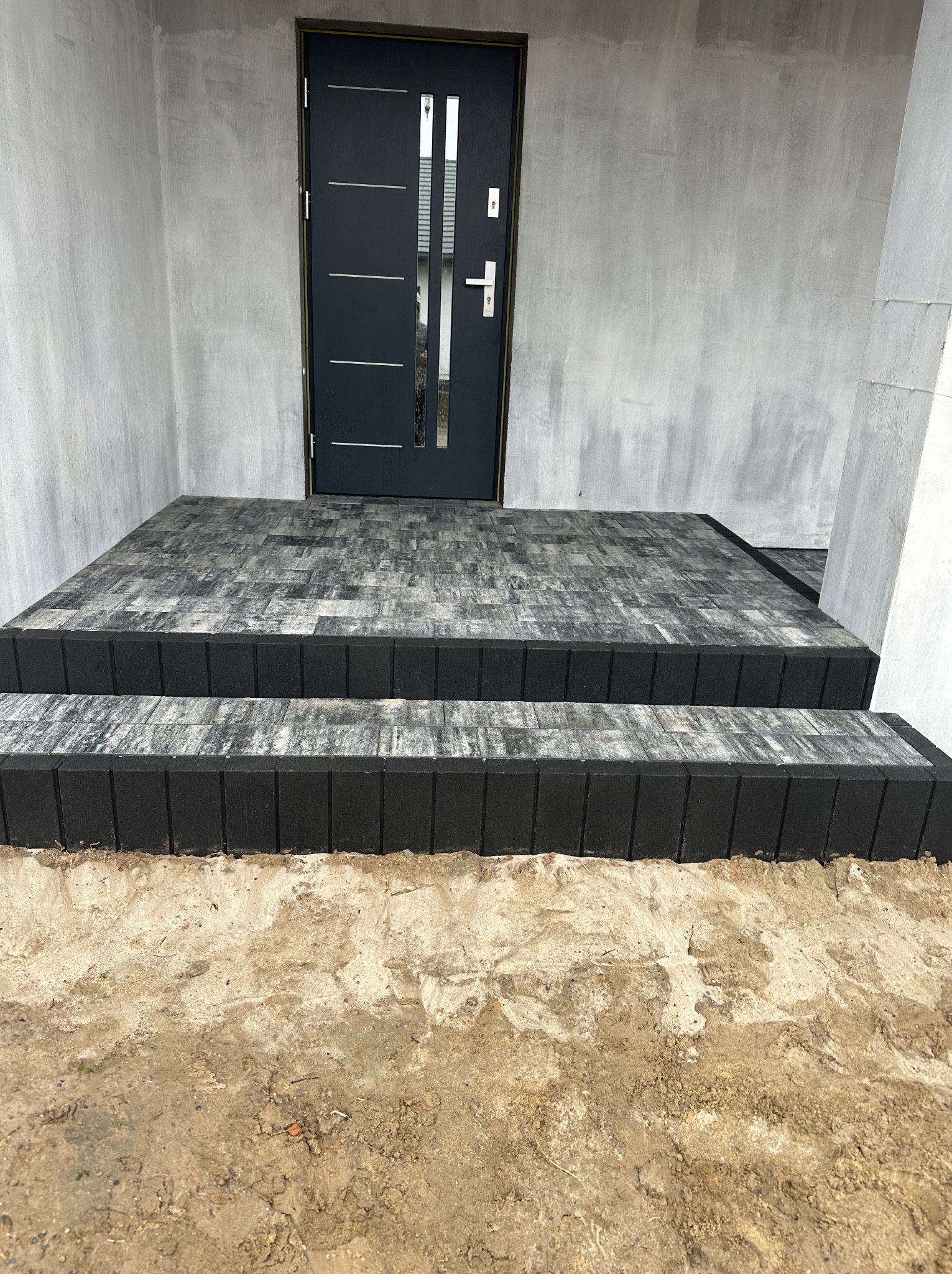 Schody wejściowe z szarej kostki brukowej z czarnym obrzeżem, prowadzące do nowoczesnych drzwi wejściowych w surowym, betonowym otoczeniu.