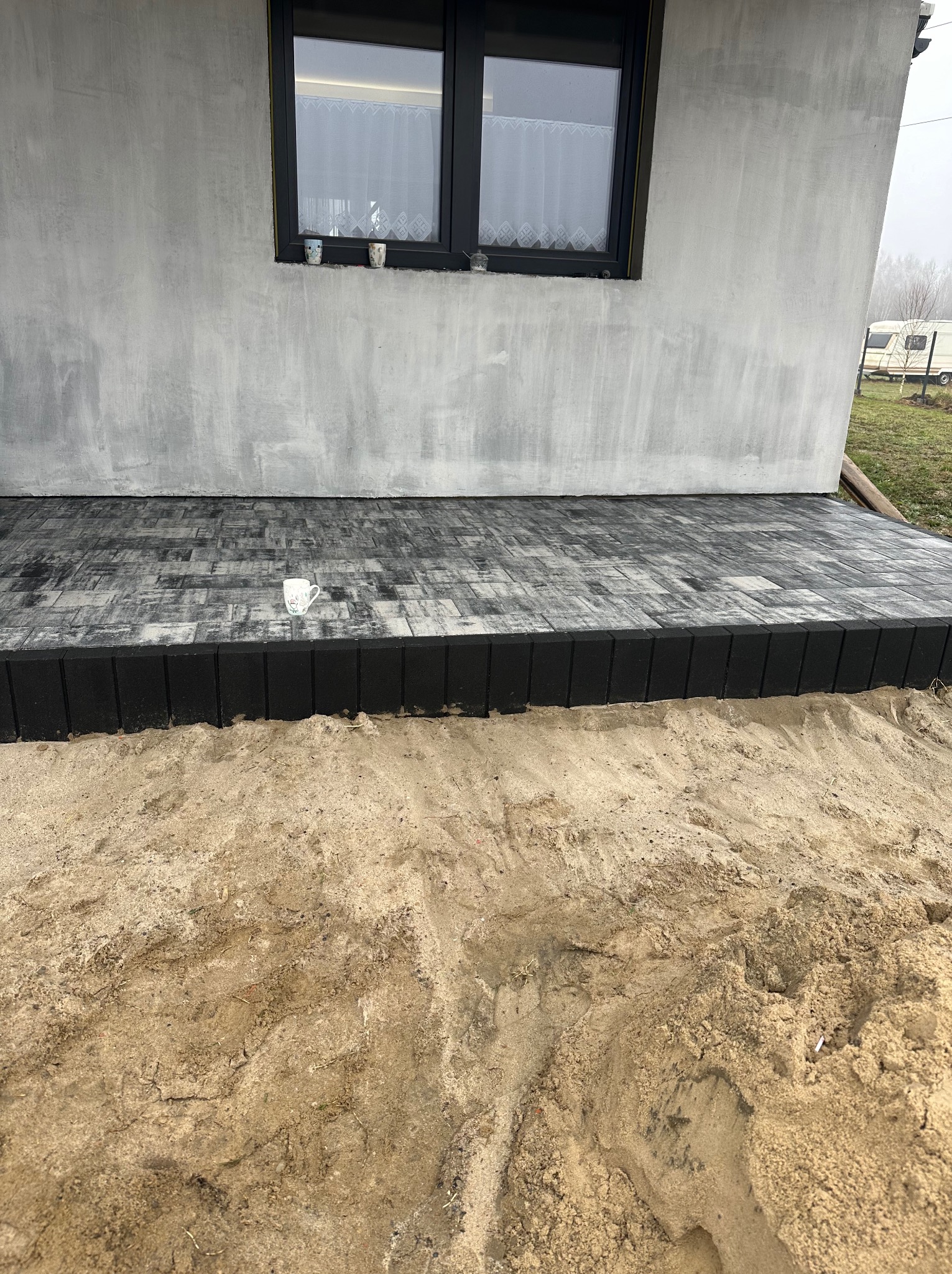 Ułożona, szara kostka brukowa tworząca taras przy szarym budynku z czarnym oknem. Na kostce stoi biały kubek. W tle piasek i zarys przyczepy kempingowej.