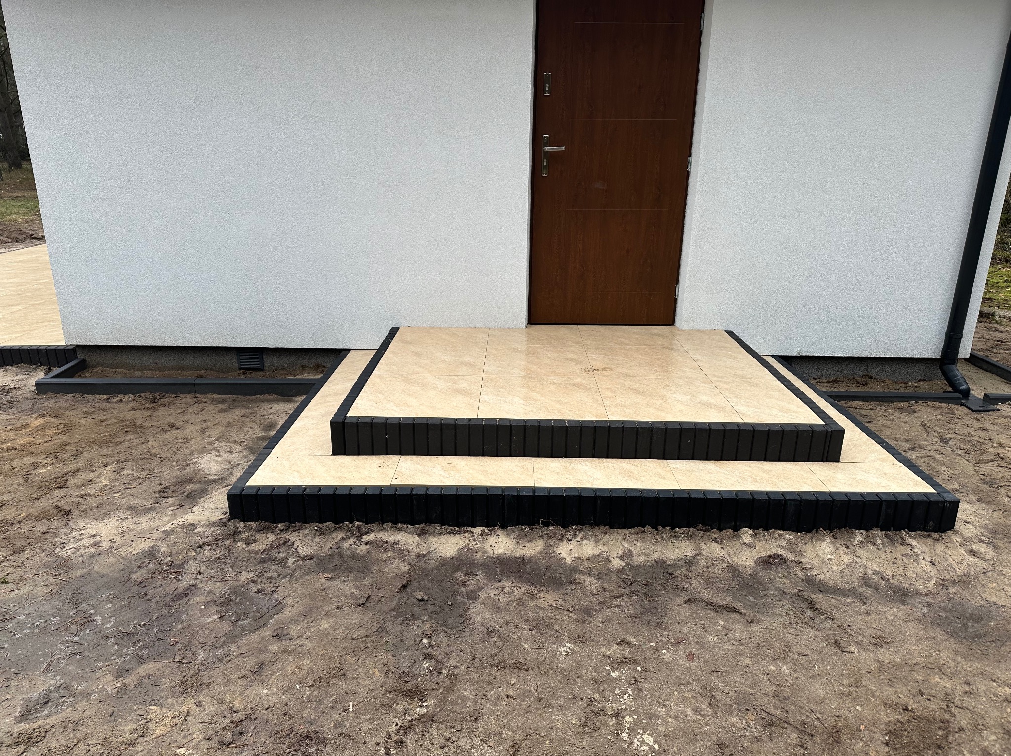 Wykończone schody wejściowe do domu, wyłożone beżową kostką brukową z czarnym obrzeżem. Widoczna elewacja budynku i fragment chodnika z kostki.